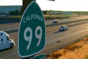 Scene along Highway 99 in California. Green road sign in foreground reading "California 99" and lanes of traffic lit by low sun.