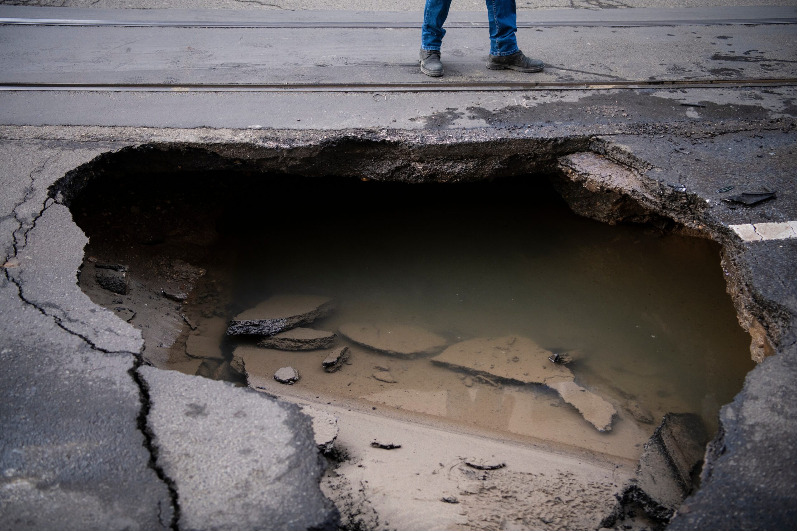 A person standing by a large sinkhole in the pavement on road. Pooled water down below.