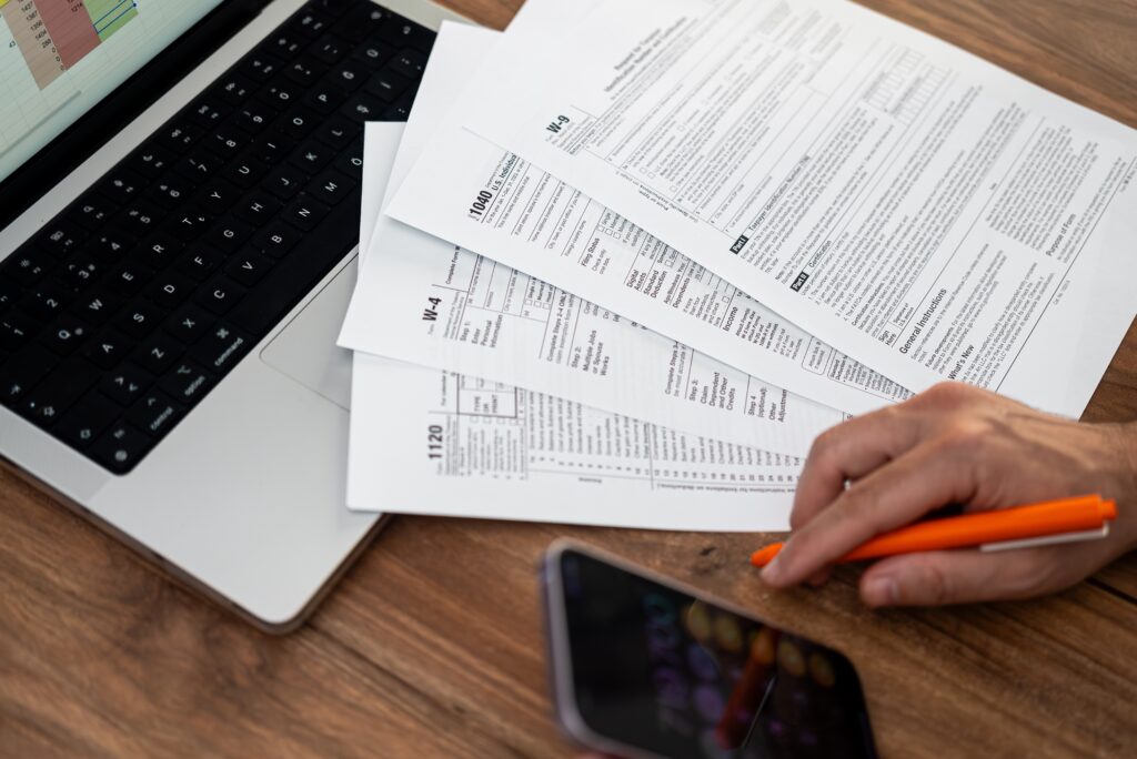 Person's hand on table holding pen with tax forms, a computer, and a phone also on table.