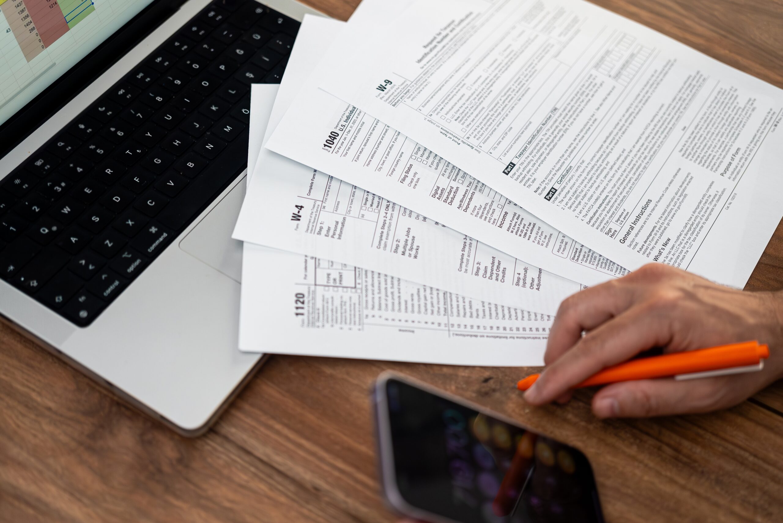 Person's hand on table holding pen with tax forms, a computer, and a phone also on table.