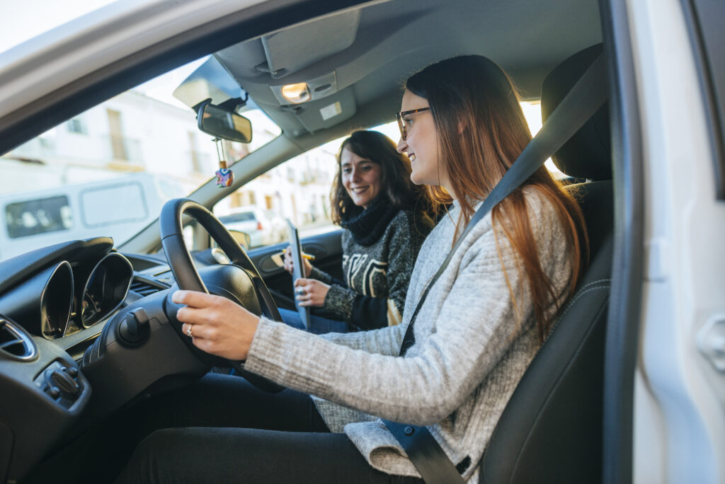 Young woman in glasses at wheel of vehicle with driving instructor sitting beside her. Driving instructor holding clipboard.