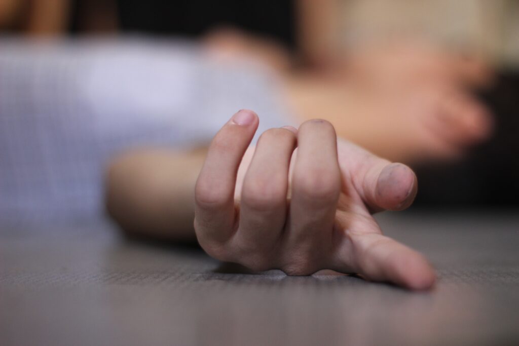 A hand in close up with a blurred man lying on the floor passed out behind.